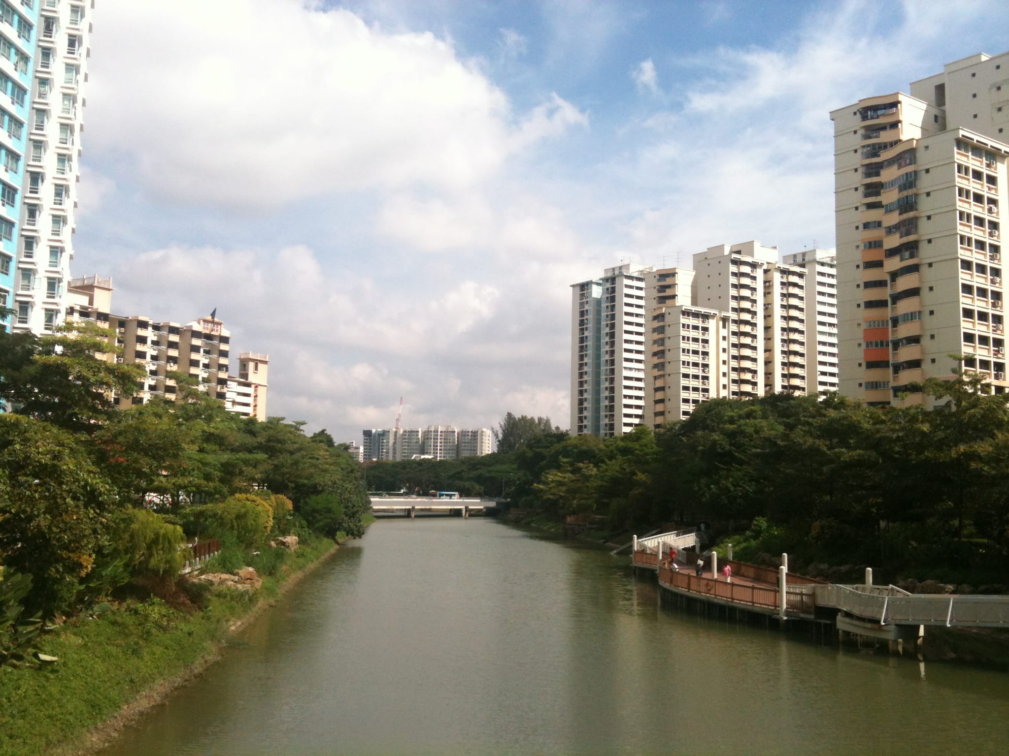 A wide river flows between tall residential buildings and a park with lush greenery on a sunny day with scattered white clouds.